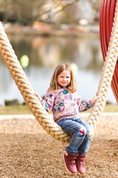 Mädchen auf einer großen Seilschaukel am Spielplatz, im Hintergrund ein Teich.
