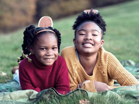 Zwei lächelnde Kinder liegen auf einer Wiese, eines in einem roten, das andere in einem gelben Pullover.