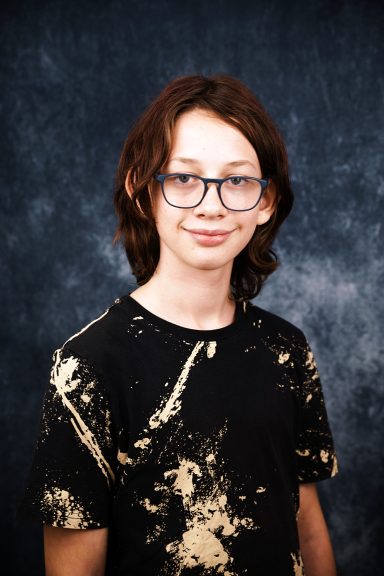 Junger Mann mit brünettem, lockigem Haar und Brille, trägt ein schwarzes T-Shirt mit Farbmustern.