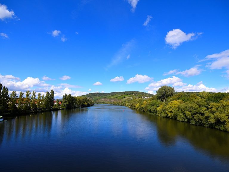 Blick auf einen ruhigen Fluss umgeben von Bäumen und blauem Himmel mit Wolken.
