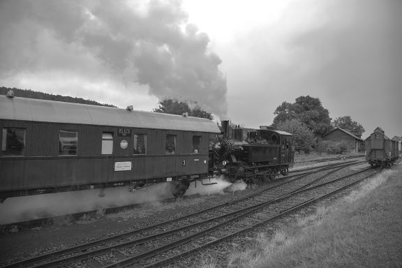 Dampflok fährt an einem Waggon vorbei, Dampfwolken steigen auf.