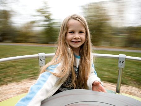 Lachendes Mädchen mit langen Haaren dreht sich auf einem Spielplatzkarussell.
