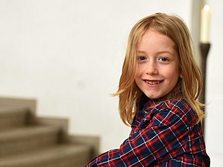 Ein lächelndes Kind mit langen blonden Haaren, trägt ein kariertes Hemd.