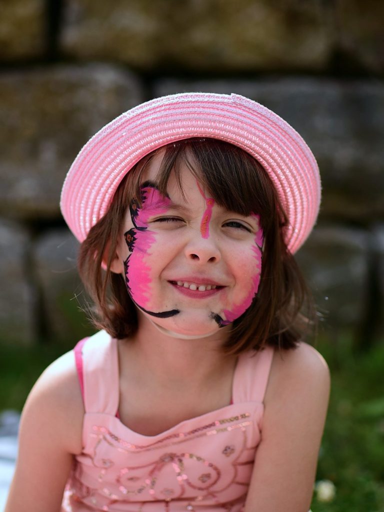 Ein Mädchen mit einem rosa Hut und geschminktem Gesicht lächelt fröhlich.